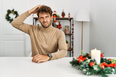 Noel süslemesi masasında oturan sakallı yakışıklı genç adam şaşırıyor ve soruyu merak ediyor. Şüpheyle kararsız, kafa kafaya düşünerek. Düşünceli kavram. 