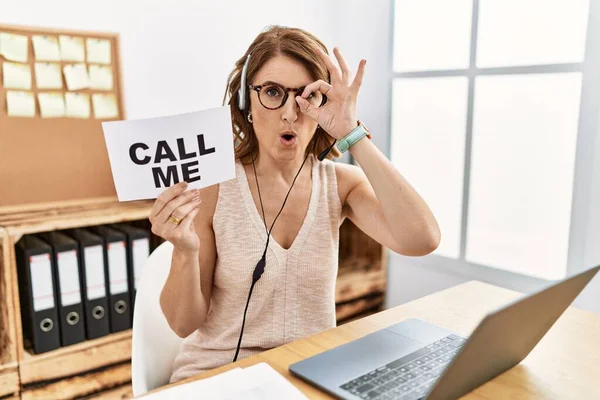 Middle age brunette woman wearing operator headset holding call me banner doing ok gesture shocked with surprised face, eye looking through fingers. unbelieving expression.