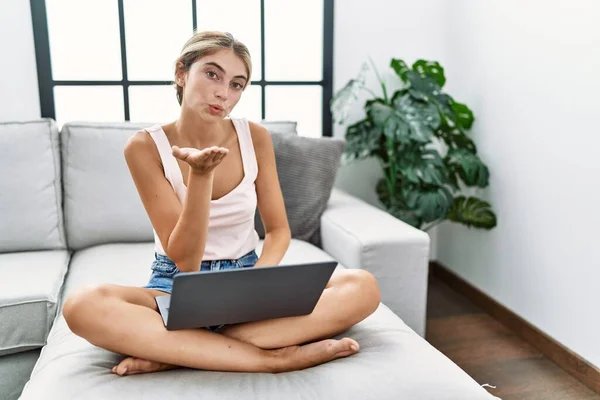 Young blonde woman using laptop at home sitting on the sofa looking at the camera blowing a kiss with hand on air being lovely and sexy. love expression.
