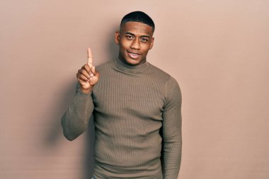 Balıkçı yaka kazak giyen genç zenci bir numaralı parmağını gösterip onu gösterirken kendinden emin ve mutlu bir şekilde gülümsüyor.. 