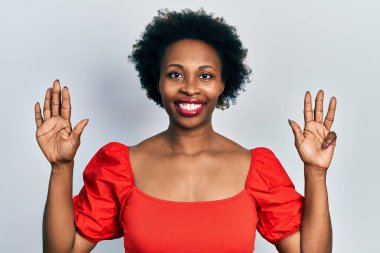 Genç Afro-Amerikalı kadın günlük giysiler giyip dokuz numaralı parmaklarıyla işaret ederken kendinden emin ve mutlu bir şekilde gülümsüyor.. 