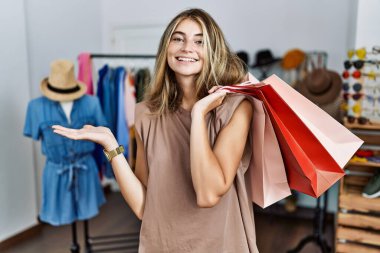 Perakende dükkanında alışveriş torbaları tutan genç sarışın kadın neşeli bir şekilde sunum yapıyor ve avuç içi kameraya bakıyor.. 