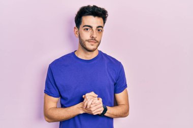 Ellerini kavuşturup gülümseyerek rahat ve neşeli bir şekilde tişört giyen genç bir İspanyol. Başarı ve iyimserlik 