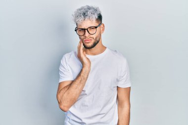 Modern boyalı saçlı, beyaz tişörtlü, gözlüklü diş ağrısı ya da diş ağrısı yüzünden ağıza dokunan, acı dolu bir ifadeyle dokunan İspanyol bir genç. dişçi 