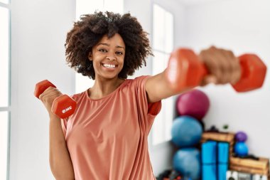 Genç Afrikalı Amerikalı kadın gülümsüyor spor merkezinde halterleri kullanarak kendine güven eğitimi alıyor