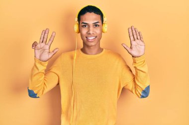 Genç Afro-Amerikalı adam müzik dinlerken kulaklık takıyor ve dokuz numaralı parmaklarıyla işaret ederken kendinden emin ve mutlu gülümsüyor.. 