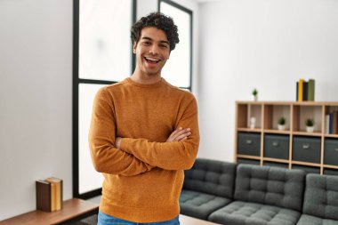 Günlük kıyafetler giyen genç İspanyol adam evde durmuş kollarını kavuşturup kameraya bakarken gülümsüyor. pozitif kişi. 