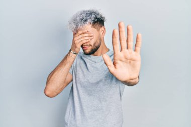 Modern boyalı saçlı, sıradan gri tişörtlü, gözlerini ellerle kapayan ve üzgün ve korku ifadeli hareketler yapan genç İspanyol bir adam. utangaç ve olumsuz kavram. 