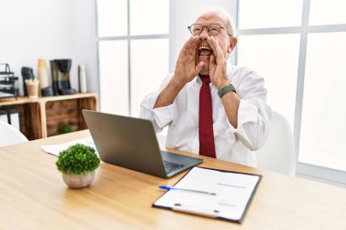 Ofiste çalışan kıdemli bir adam bilgisayar kullanıyor ve elleri ağızda bağırıyor. 