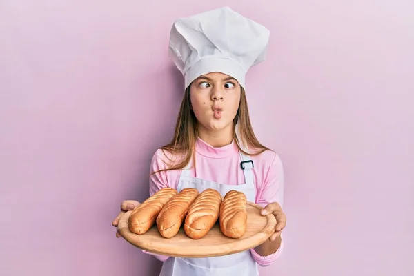 Beautiful brunette little girl wearing baker uniform holding homemade bread making fish face with mouth and squinting eyes, crazy and comical.