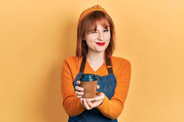 红头发的年轻女子手持女服务员围裙 一边喝着咖啡 一边眨着眼睛 带着性感的表情 笑容满面地看着相机 — 图库照片