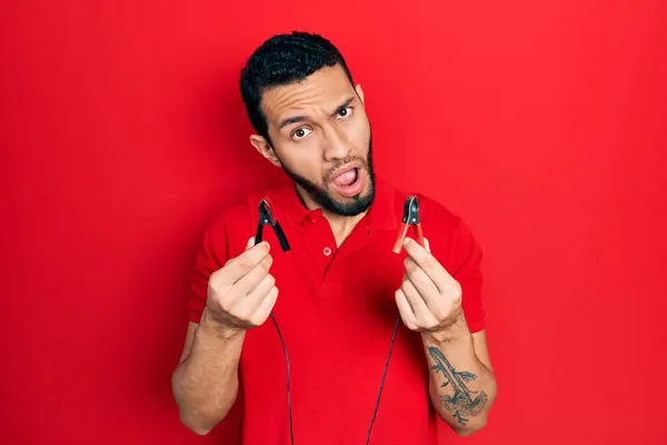 Hispanic man with beard holding battery clamps in shock face, looking skeptical and sarcastic, surprised with open mouth