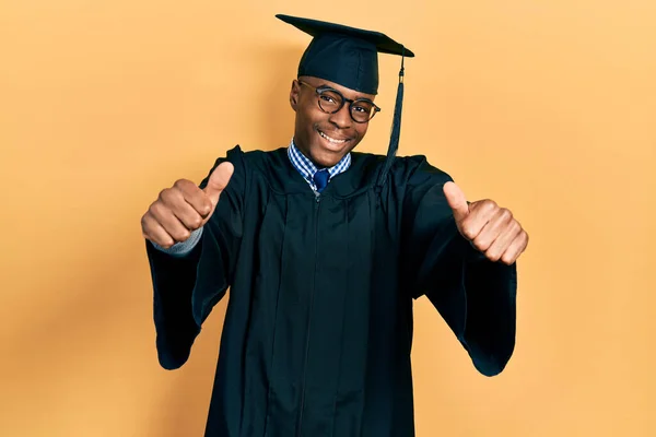 卒業キャップと式のローブを着た若いアフリカ系アメリカ人男性は 手で積極的なジェスチャーを行うことを承認し 親指を上げて笑顔と成功のために幸せ 勝者のジェスチャー — ストック写真
