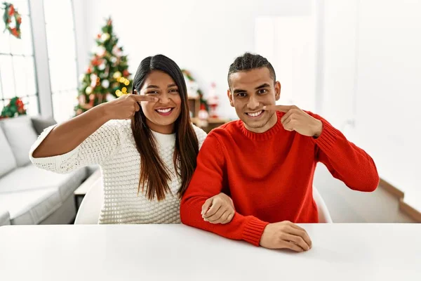 Young Latin Couple Sitting Table Christmas Tree Pointing Hand Finger — Stockfoto