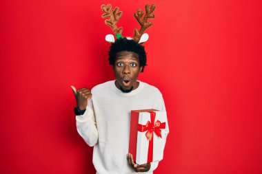 Geyik Noel şapkası takan genç Afro-Amerikan bir adam elinde hediyeyle elini yana uzatarak şaşırdı, ağzı açık hayret içinde kaldı.. 