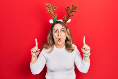 Orta yaşlı beyaz kadın şirin noel geyiği boynuzları takıyor şaşırmış ve şaşırmış görünüyor ve parmaklarıyla işaret ediyor ve kollarını kaldırıyor.. 