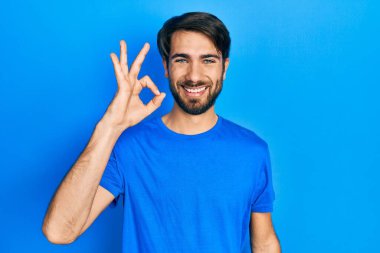 Gündelik kıyafetler giyen genç İspanyol adam gülümsüyor. El ve parmaklarıyla 