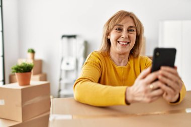 Orta yaşlı sarışın kadın yeni evinde akıllı telefon kullanarak mutlu bir şekilde gülümsüyor..