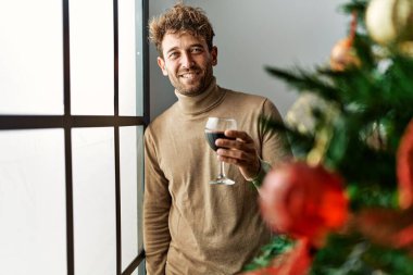 Genç İspanyol adam evde Noel ağacının yanında şarap içiyor.