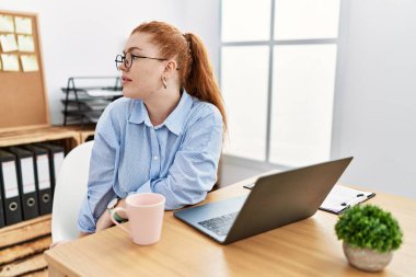 Ofiste çalışan genç kızıl saçlı bir kadın bilgisayar kullanıyor. Yan tarafa bakıyor, profil pozu veriyor. Doğal bir yüzü ve kendine güvenen bir gülümsemesi var.. 