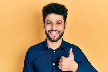 Sakallı, sıradan gömlekli genç bir Arap, mutlu bir el hareketi yapıyor. Kameraya bakıp başarıyı gösteren ifade. 