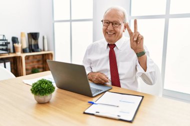 Ofiste çalışan kıdemli bir adam bilgisayarla iki numarayı gösterip işaret ederken kendinden emin ve mutlu bir şekilde gülümsüyor.. 
