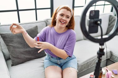 Genç kızıl saçlı kadın evde akıllı telefon kullanarak makyaj dersi veriyor.