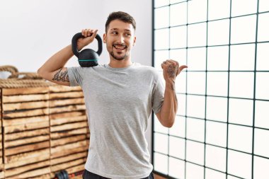 Spor kıyafetleri giyen genç yakışıklı adam halterleri kullanarak baş parmağını kaldırıyor ve ağzı açık gülümsüyor. 