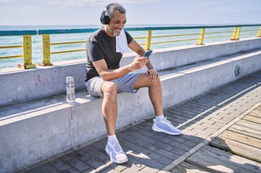 Orta yaşlı İspanyol adam deniz kenarında akıllı telefon kullanarak spor giyer.