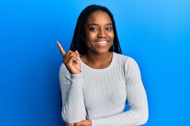 Saçları örgülü beyaz süveter giyen, yüzünde kocaman bir gülümseme olan, elleri ve parmaklarıyla kameraya bakan Afro-Amerikan bir kadın.. 