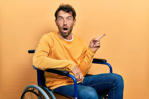 Handsome man with beard sitting on wheelchair surprised pointing with finger to the side, open mouth amazed expression.