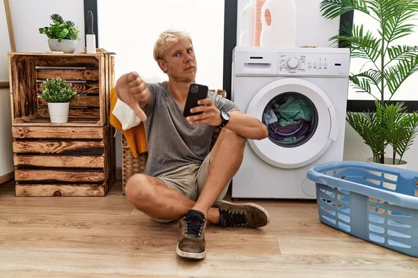 Genç Sarışın Adam Çamaşır Yıkıyor Akıllı Telefon Kullanıyor Mutsuz Görünüyor — Stok fotoğraf