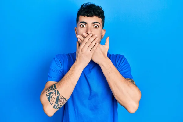 カジュアルなブルーのTシャツを着た若いヒスパニック系の男性がミスのために手で口をカバーショックを受けた 秘密の概念 — ストック写真