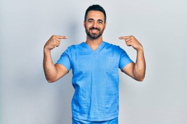 Sakallı yakışıklı, mavi erkek hemşire üniforması giyen, kendine güvenen, kendini gururlu ve mutlu parmaklarıyla gösteren bir İspanyol.. 