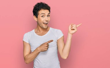 Gündelik beyaz tişört giyen genç yakışıklı adam gülümseyip kameraya bakıyor ve iki eliyle ve parmaklarıyla onu işaret ediyor.. 