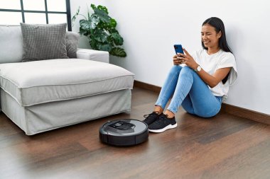 Evdeki akıllı robot süpürgesiyle yerleri temizleyen genç Latin kadın.