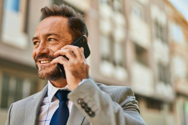 Orta yaşlı işadamı gülümsüyor. Şehirdeki akıllı telefondan konuşuyor..