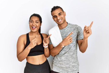 Young latin couple wearing sportswear standing over isolated background smiling and looking at the camera pointing with two hands and fingers to the side. 