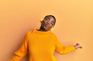 Kısa saçlı, kulaklık takan, müzik dinleyen ve izole edilmiş sarı arka planda dans eden güzel Afrikalı Amerikalı genç kadın.