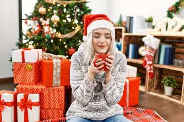 Beyaz kız mutlu bir şekilde gülümsüyor Noel şapkası takıp evde kahve içiyor..