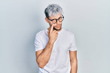 Modern boyalı saçlı, beyaz tişörtlü ve gözlüklü genç İspanyol adam senin jestini izlerken şüpheli bir ifade takınıyor. 
