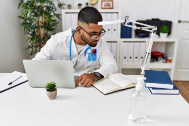 Klinikte doktor üniforması giyen genç bir ortadoğu adamı kitap okuyor.