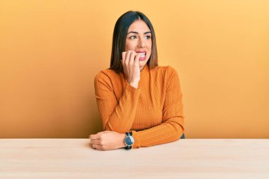 Gündelik kıyafetler giyen genç İspanyol kadın masada oturmuş gergin ve gergin görünüyor. Ağzı tırnak yiyen ellerle. anksiyete. 