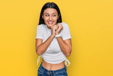 Latin kökenli genç bir kız, günlük beyaz tişört giyiyor. Gergin ve heyecanlı bir şekilde gülüyor. Elleri çenesinde yan tarafa bakıyor. 