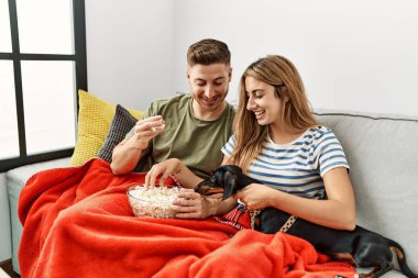 Genç İspanyol çift gülümsüyor. Mutlu bir şekilde kanepede oturmuş, evde köpekleriyle film izliyorlar..