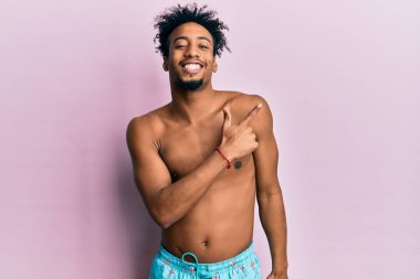 Yüzünde mutlu ve doğal bir ifade olan, elleri ve parmaklarıyla gülümseyen, mayo giymiş, sakallı, Afro-Amerikalı genç bir adam. 