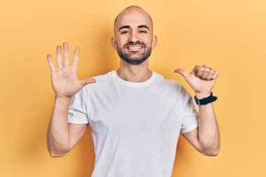 Genç kel adam, günlük beyaz t-shirt giyip altı numaralı parmaklarıyla işaret ederken kendinden emin ve mutlu bir şekilde gülümsüyor.. 