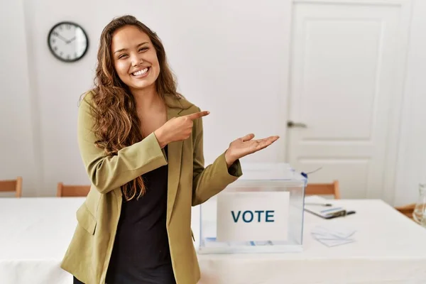 Beautiful hispanic woman standing at political campaign room amazed and smiling to the camera while presenting with hand and pointing with finger.