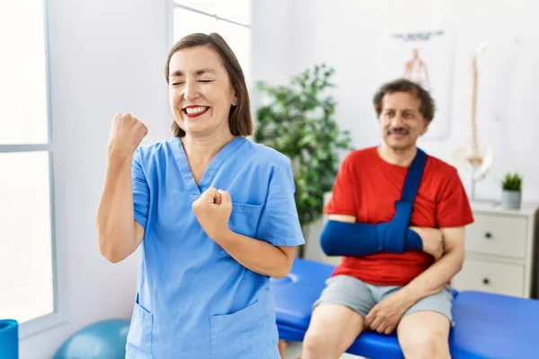 Middle age doctor woman with patient with arm injury at rehabilitation clinic celebrating surprised and amazed for success with arms raised and eyes closed
