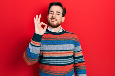 Sakallı, zarif yün süveter giyen yakışıklı beyaz adam gülümsüyor ve el ve parmaklarıyla imza atıyor. başarılı ifade. 